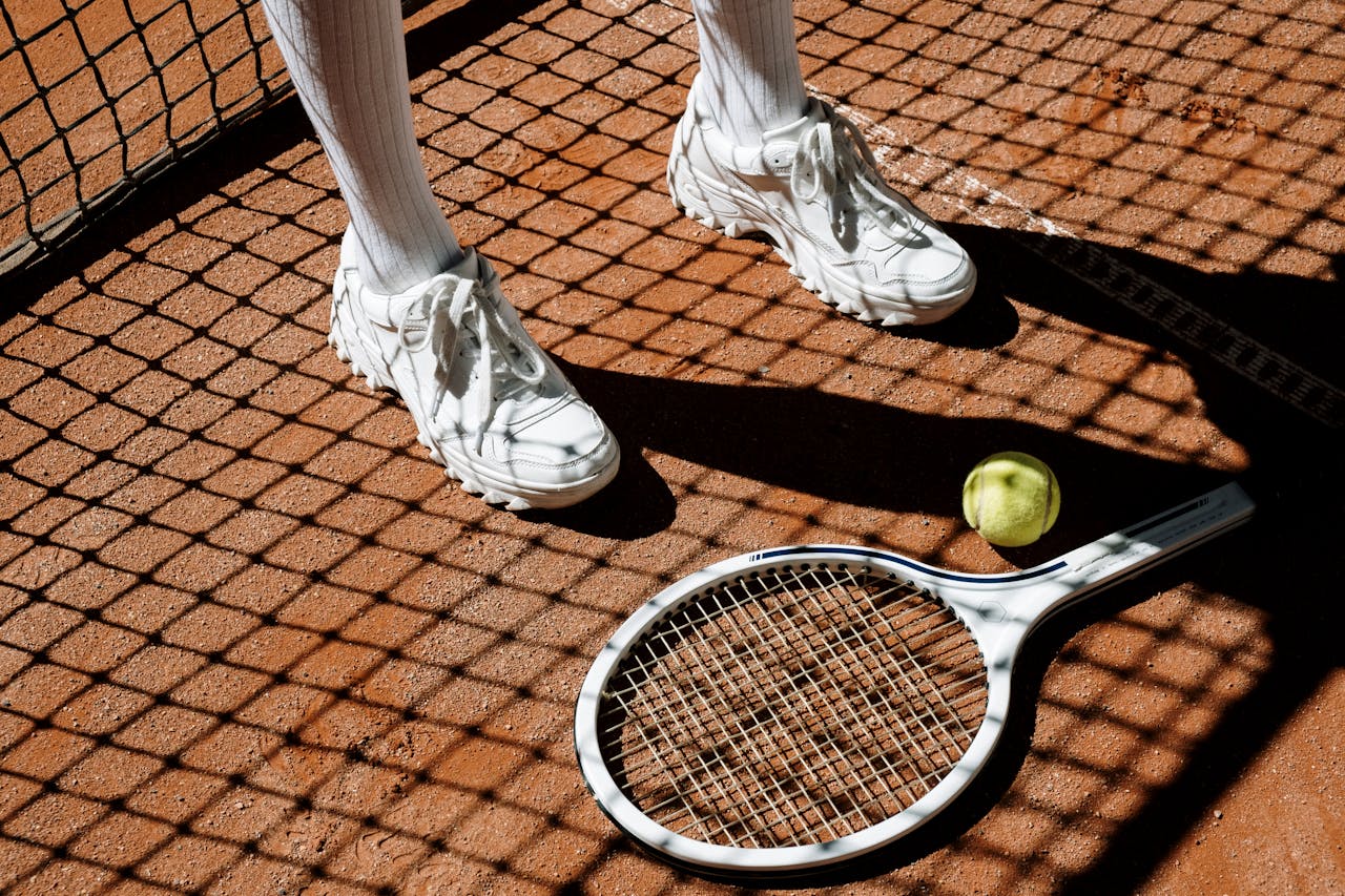 Tennis players legs with racket and ball on clay court under bright sun and shadow patterns.